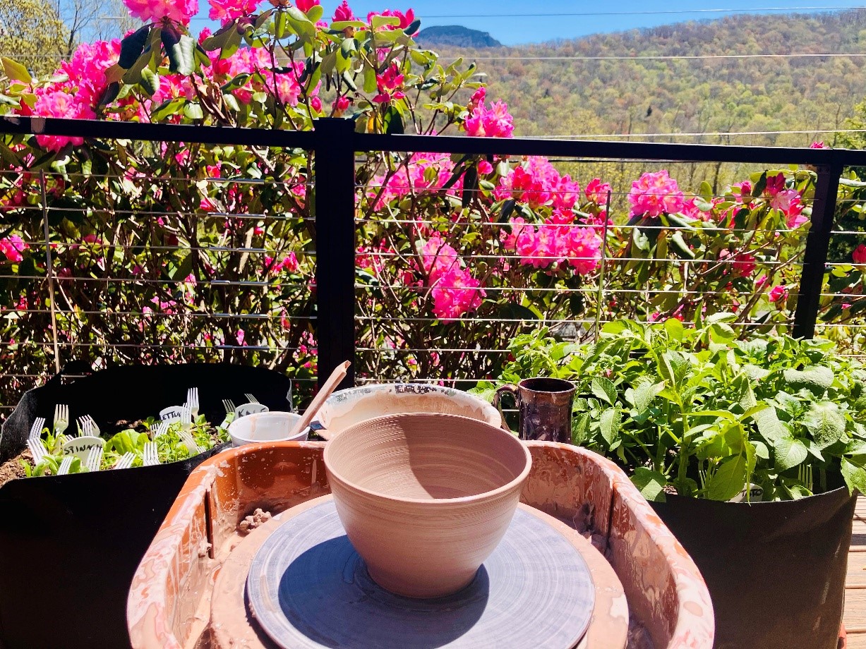 A clay bowl being shaped on a pottery wheel on a mountain deck with bright spring flowers and ridges in the background.
